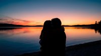 Romantic couple silhouette embracing at sunset by calm lake with colorful sky reflection on water creating peaceful intimate moment for love and relationship concepts.