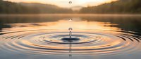 Water drop creating ripples on calm lake surface at golden hour sunset with mountains in background creating peaceful meditation scene.