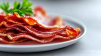 Crispy fried bacon strips garnished with fresh parsley on white ceramic plate, close-up view with selective focus showing texture and shine of cooked meat.