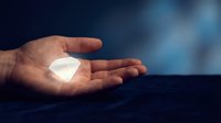 Hand holding glowing white geometric crystal or gem stone against dark blue blurred background. Concept for innovation, technology, energy, and future solutions.