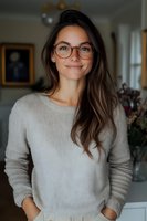 Young Caucasian woman with long brown hair wearing glasses and beige sweater smiling warmly at camera. Natural indoor lighting creates soft ambiance for lifestyle portrait.