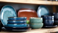 Stacked ceramic plates and bowls in turquoise, blue and brown colors on wooden shelf. Handcrafted pottery dinnerware collection showcasing artisanal glazing techniques.