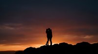 Romantic couple silhouette embracing on mountain peak during golden sunset with dramatic orange and purple sky creating intimate moment for love concepts.
