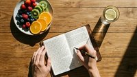 Overhead view of person reading open book at wooden table with fresh fruit plate, orange slices, berries, and water glass. Relaxing reading moment.