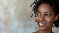 Young African American woman with natural dreadlocks hairstyle and bright white smile against blurred background, radiating confidence and joy in casual portrait.