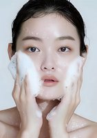 Young Asian woman applying facial cleanser foam, demonstrating skincare routine with fresh glowing complexion against white background for beauty and wellness concept.