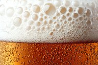 Fresh beer with white foam and bubbles in glass, extreme close up macro photography showing amber liquid and carbonation texture against light background.