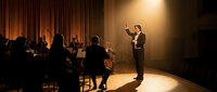 Professional orchestra conductor leading symphony performance on illuminated concert hall stage with musicians and warm theatrical lighting.