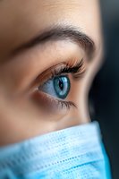 Close-up view of bright blue eye with long eyelashes and defined eyebrow above protective medical face mask, representing healthcare and safety during pandemic.