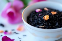 Loose black tea leaves in white ceramic bowl with scattered pink rose petals and flower buds, selective focus on orange petal against blurred floral background.