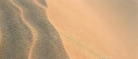 Young adult male with light skin tone wearing casual beige clothing, reclining on sandy beach with warm golden sunlight creating soft shadows across textured sand surface.