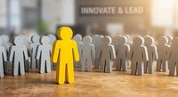 Yellow figure stands out from crowd of white paper cutouts on wooden surface with innovate and lead text in blurred office background for leadership concepts.