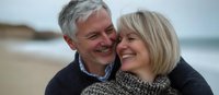 Mature Caucasian couple sharing intimate moment at beach, laughing together with genuine joy. Gray-haired man embraces blonde woman wearing knit scarf on cloudy day.