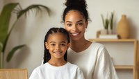 African American mother and daughter smiling at home, wearing white casual clothing. Natural light interior with houseplants and modern minimalist decor in background.