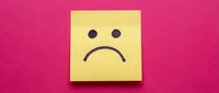Yellow sticky note with sad face drawn on pink background expressing disappointment and negative emotions for mental health awareness campaigns.