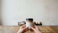 Hands holding blank white coffee cup with black lid on wooden table, suitable for branding and cafe marketing materials.