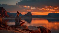 Young woman practicing meditation yoga pose on rocky cliff overlooking serene lake at golden sunset with dramatic orange sky clouds reflecting in water.