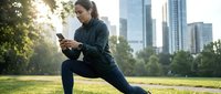 Young woman of mixed ethnicity in athletic wear performing a lunge stretch in urban park with skyline, using smartphone for fitness tracking or workout app during outdoor exercise session.
