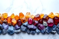 Raw colorful gemstones arranged in gradient pattern from yellow to red to blue on white marble background, showing natural crystal formations and mineral textures.