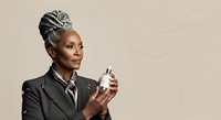 Mature African American woman with braided updo hairstyle wearing black blazer holding fragrance bottle against neutral background for beauty and cosmetics marketing.