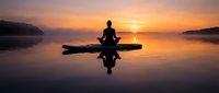 Silhouette of person meditating in lotus pose on paddleboard at sunset over calm lake with perfect reflection creating peaceful mindfulness scene.