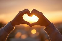 Hands forming heart shape silhouette against golden sunset sky with warm bokeh lights creating romantic atmosphere for love and affection concepts.
