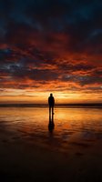 Silhouetted male figure standing on wet beach sand at dramatic sunset with golden and orange sky reflecting on water.