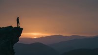 Young adult male silhouette standing on rocky cliff edge at sunset overlooking mountain landscape. Concept for achievement, adventure, success, and personal growth in outdoor settings.