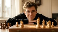 Young Caucasian man with glasses and curly hair playing chess at wooden board in cozy indoor setting with warm natural lighting from window.