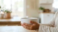 Closeup of hands holding warm ceramic mug in bright kitchen with blurred background, suitable for beverage, lifestyle, and wellness marketing materials.