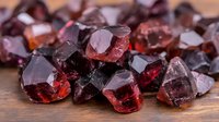 Raw garnet crystals and stones in deep red color scattered on wooden surface, showing natural mineral formation with rough edges and translucent properties.