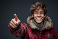 Young Caucasian man in red plaid winter jacket with fur hood and gray sweater showing thumbs up gesture while smiling cheerfully against dark background.