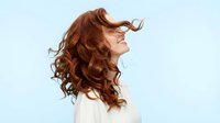 Young woman with flowing red curly hair in motion against light blue background expressing joy and freedom for wellness and beauty concepts.