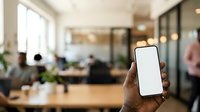 Cropped hand holding smartphone with blank white screen in modern office workspace with colleagues working at desks, plants, and natural light for app mockup and business communication.