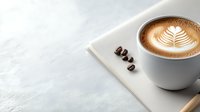 Cup of cappuccino with latte art fern pattern on white surface, coffee beans scattered nearby. Minimalist composition with soft natural lighting and copy space.