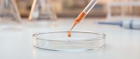 Laboratory pipette dropping orange liquid sample into glass petri dish for scientific research and medical testing in modern lab setting.