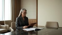 Mature Caucasian businesswoman with gray hair and glasses reviewing documents at conference table in modern office boardroom for corporate meetings.