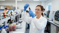 Young female scientist with brown hair and fair complexion wearing white lab coat and blue latex gloves examining test tube in modern laboratory setting with equipment and samples.