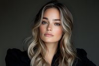Young woman with blonde balayage hair and natural makeup poses against dark background, wearing black top. Professional beauty portrait with soft lighting.