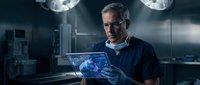 Senior male surgeon with gray hair and glasses examining digital holographic tablet in modern operating room with surgical lights and medical equipment.