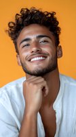 Young Latino man with curly hair and bright smile wearing white shirt against vibrant orange background, hand thoughtfully placed on chin in confident pose.