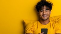 Young Asian man with bright smile using smartphone while relaxing on yellow background, expressing joy and positive emotions during mobile browsing.