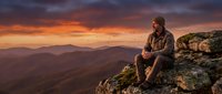 Contemplative hiker sits on rocky mountain summit during golden hour sunset with dramatic orange sky and distant peaks for outdoor adventure concepts.