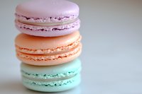 Pastel colored French macarons stacked in pink, peach and mint green shades against soft blurred background, showcasing delicate meringue texture and cream filling.