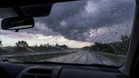 Stormy weather driving view through car windshield with raindrops on glass and dark clouds ahead on empty rural road during severe weather conditions.