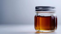 Glass jar filled with golden amber liquid against soft gray background, minimalist composition highlighting transparency and warm honey-like tones.