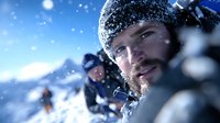 Young bearded man in winter hat with frost on facial hair against snowy mountain backdrop during outdoor adventure with friends in background.