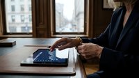 Mature woman in business attire working on tablet at wooden desk with city view through windows, professional office environment for corporate and technology concepts.