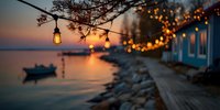 Cozy lakeside cottage with string lights and lanterns at sunset, wooden deck overlooking calm water with small boat silhouette.