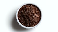 Ground dark cocoa powder in white ceramic bowl on white background, overhead view showing rich brown color and fine powder texture. Isolated food ingredient.